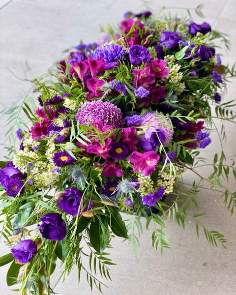 A floral casket spray with a mix of purple and pink flowers, green leaves, and decorative elements.