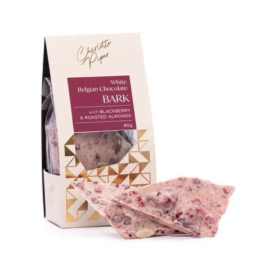 A piece of Charlotte Piper White Chocolate Belgian Bark with roasted almonds visible, placed in front of its packaging box.