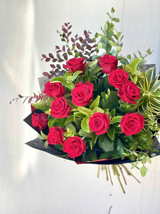A bouquet of red roses with green leaves and some berries, wrapped in a paper wrapper.