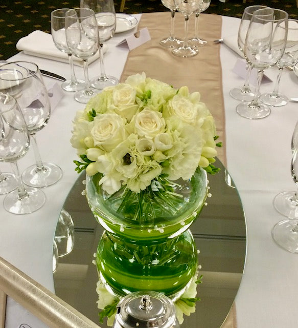 A floral centrepiece with white blooms including roses, freesia and lissianthus in a modern round glass vase with green detailing, placed on a table set for an event.