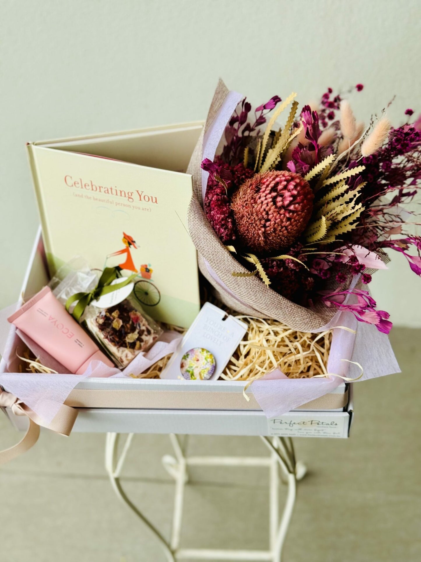 A gift hamper containing a bouquet of preserved flowers, a scented candle, a book titled 'CELEBRATING YOU', hand lotion, nougat, and a fridge magnet, all presented in a beige gift box with pink accents.