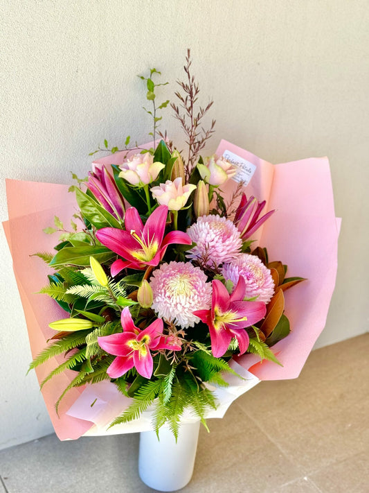 A bouquet of flowers featuring shades of pink including oriental lilies, roses, and disbud chrysanthemums, wrapped in pink paper.