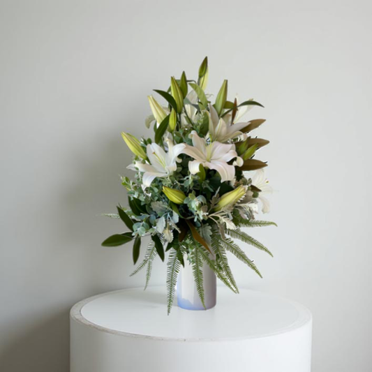 A white ceramic vase filled with white oriental lilies and green foliage, displayed on a white pedestal against a neutral background.