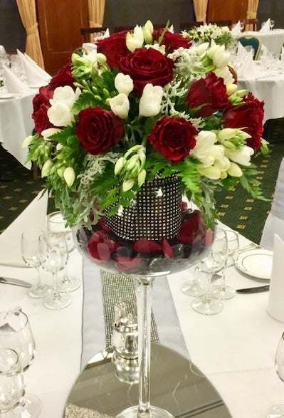 A floral arrangement in a Bordeaux-style wine glass with a sparkling base, featuring red and white flowers including roses and freesias, designed as a centrepiece for a table setting.