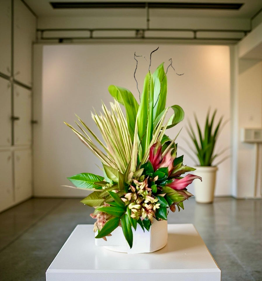 A greenery arrangement featuring a mix of bold green leaves and tropical accents, arranged in a white pot, displayed in a bright indoor space. An ideal centrepiece for an office environment that requires longevity.