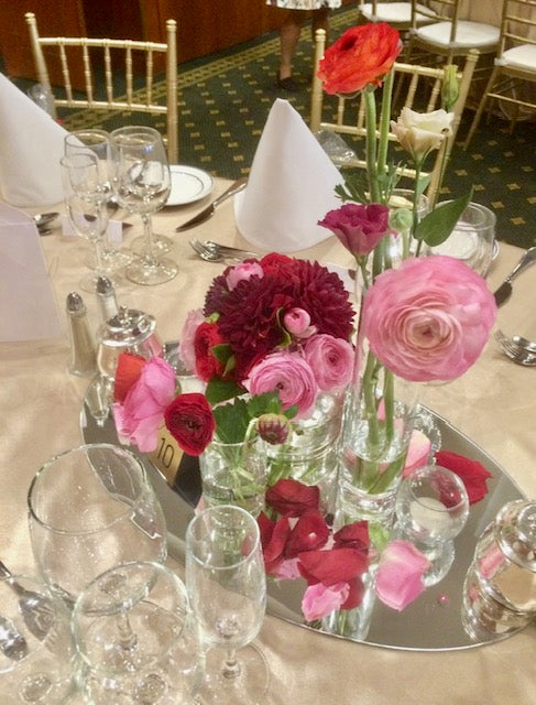 A table setting with a stunning floral arrangement at the center, including red and pink flowers including lissianthus and dahlias, complemented by individual bud vases and scattered rose petals.