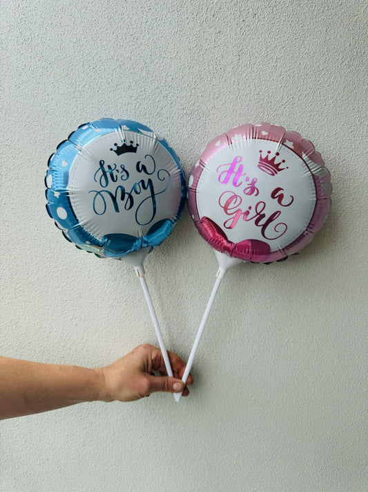 Two round balloons, one pink and one blue, with 'It's a boy' and 'It's a girl' text along with a crown design, held by a hand against a neutral background.
