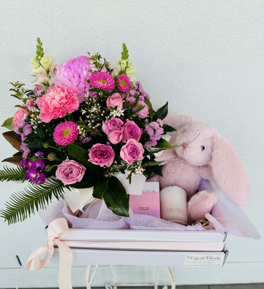 A baby hamper gift set featuring a basket with seasonal flowers in soft pink and white hues, a plush toy Nana Huchy rabbit, and a mini Ecoya Sweet Pea & Jasmine candle, all tied together with a pink ribbon.