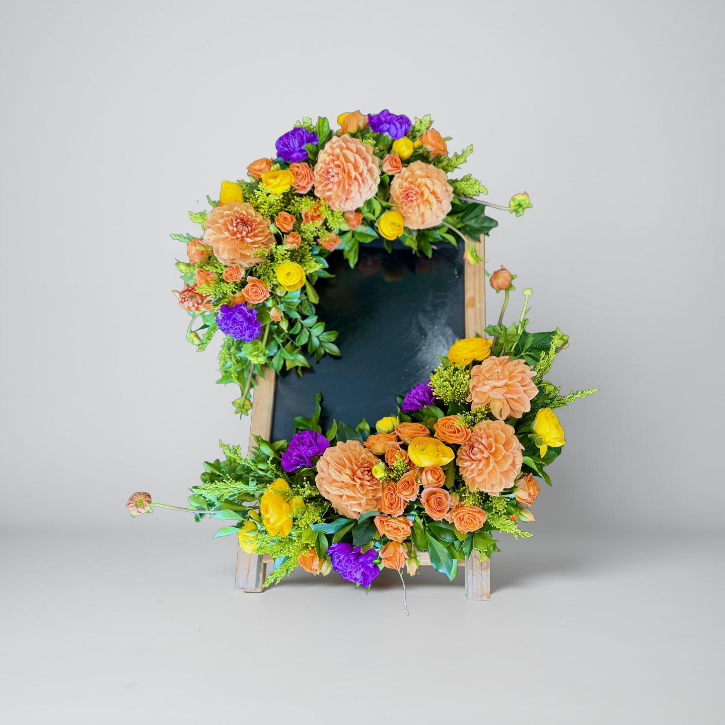 Celebration Floral Frame with vibrant fresh flowers in orange, yellow, purple, and peach arranged around a large wooden event sign.