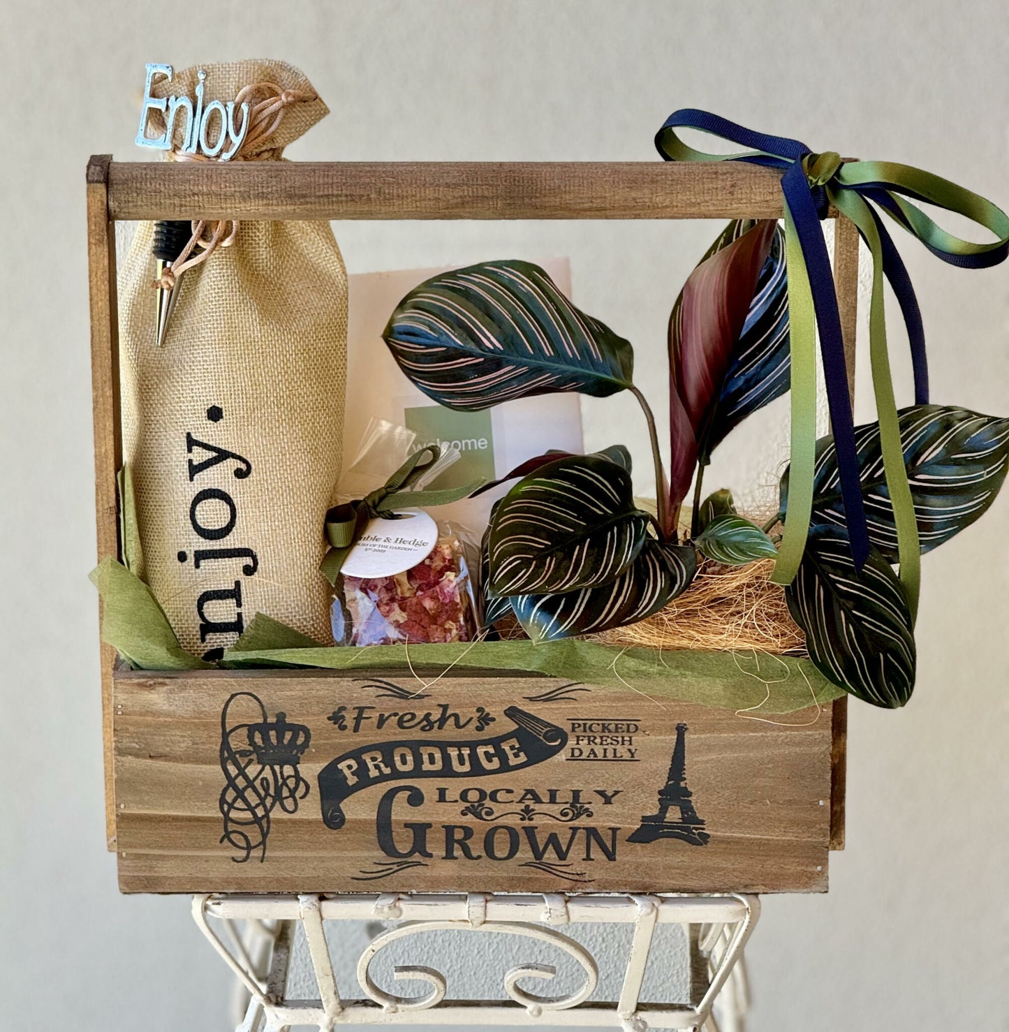 A rustic wooden crate with the word 'Fresh Produce Locally Grown' printed on it, containing a bottle of wine in a hessian bag marked 'Enjoy" with a matching wine stopper, a green plant, a book titled 'Welcome', and a small nougat treat.