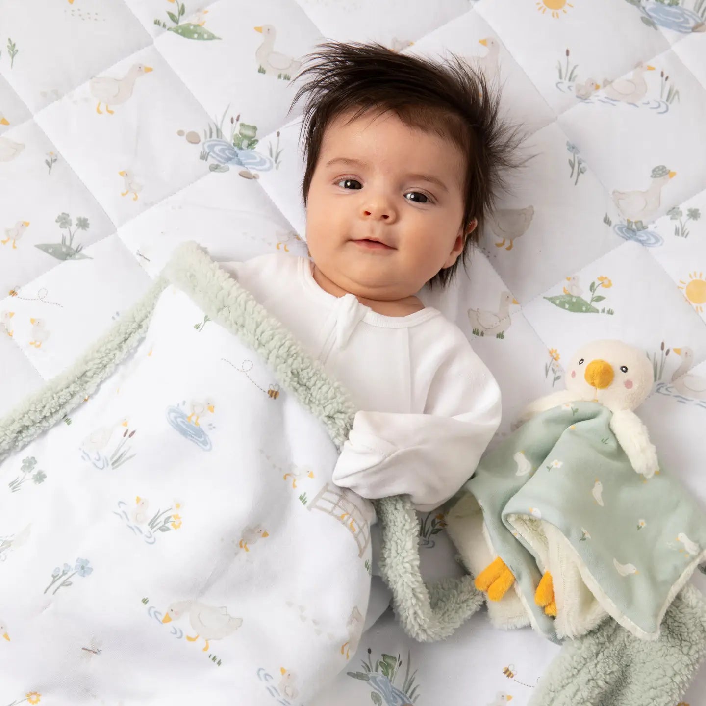 A baby lying on a pastel green security blanket with a pattern of geese and flowers, holding a plush duckling.