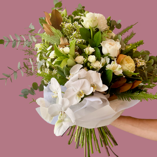 An elegant floral bouquet featuring a mix of white roses, lush orchids, and green foliage, wrapped in white paper and ribbon.
