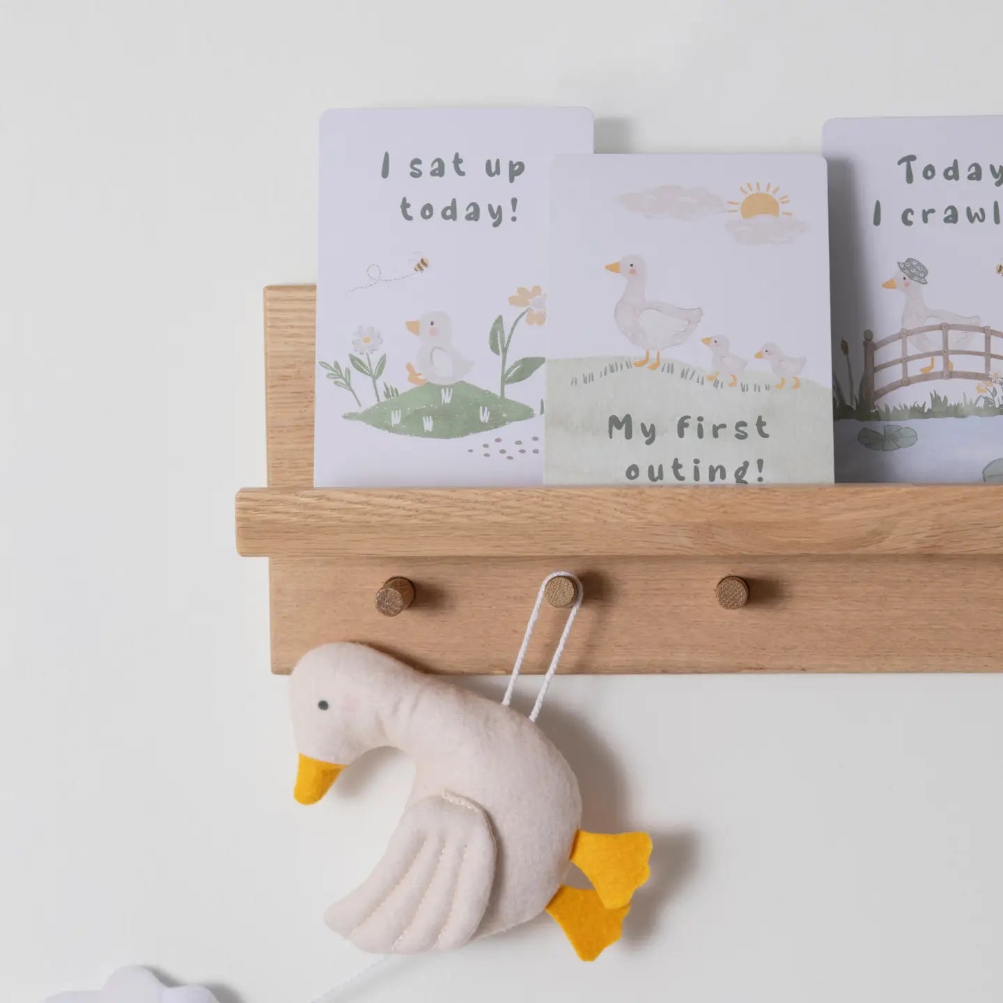 A set of illustrated baby milestone cards featuring a goose and her baby, displayed on a wooden shelf with a plush toy goose below.