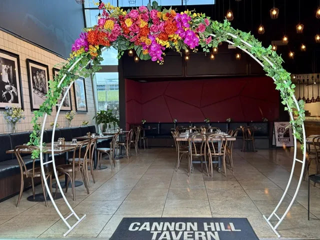 A circular white arch dressed with bright pink, orange and green flowers and foliage, framing the entrance to a venue