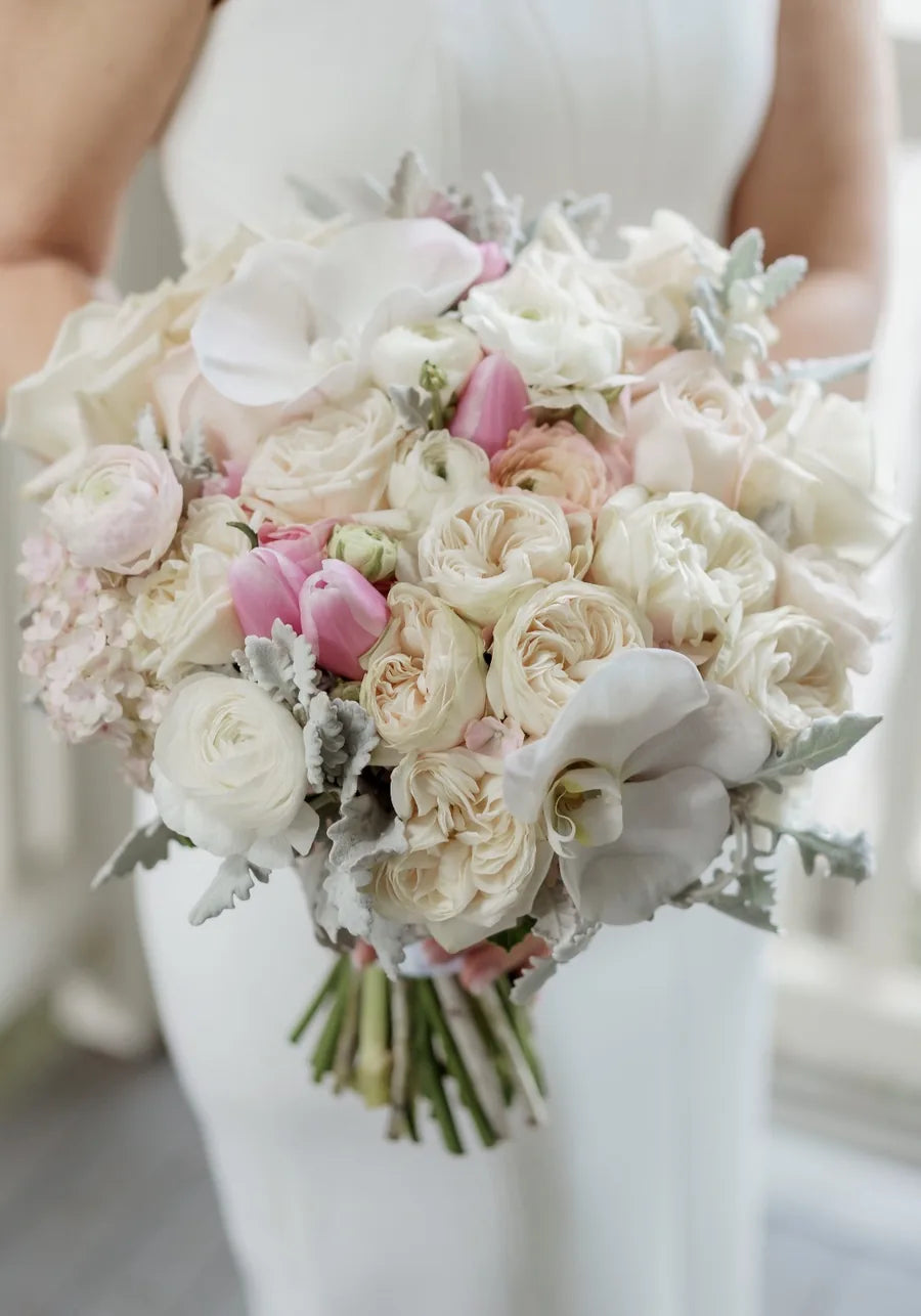 A bride in a white gown holding a soft blush and ivory bouquet of garden roses, ranunculus, tulips and phalaenopsis orchids