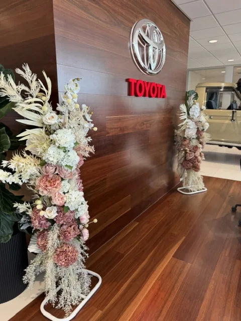 Two floral pillars in blush, cream and dried tones standing against a timber feature wall in a corporate showroom foyer