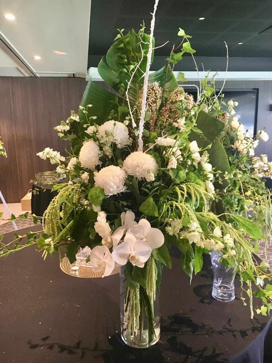 A photograph displaying three luxurious floral arrangements with a predominance of white flowers and green foliage, arranged in clear vases, intended for event decor.