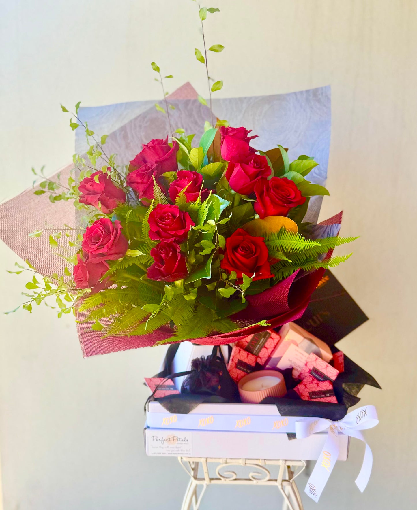 A gift arrangement that includes a dozen red roses, scattered rose petals, a scented candle, a bottle of fine wine, and indulgent chocolates presented in a box.