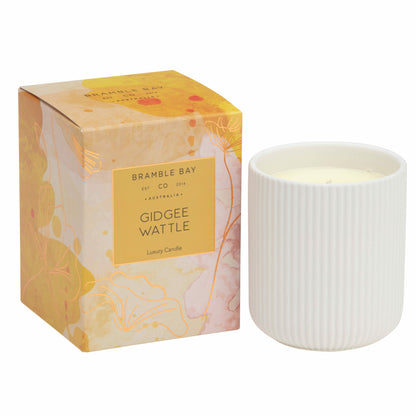 A Bramble Bay Co Gidgee Wattle white ceramic candle with a coppery rose foil lid, labeled 'Gidgee Wattle' by Bramble Bay Co. with 'Australian Native' fragrance, displayed next to its box with abstract Australian floral artwork.