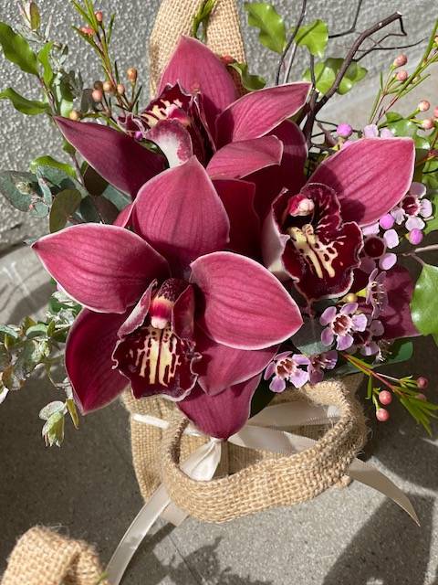 A hessian bag arrangement featuring various cymbidium orchids in shades of pink and purple, tied with twine and set against a burlap background.