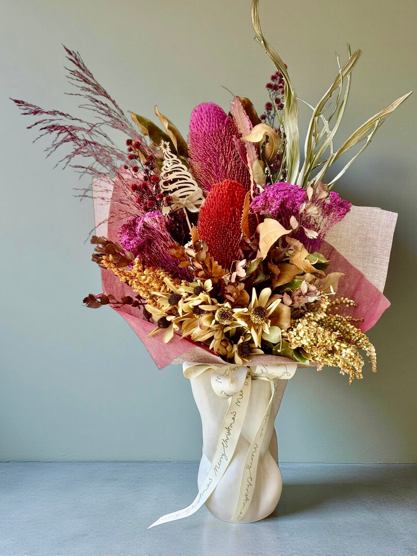 Australian summer Christmas dried flower arrangement in festive red and gold tones with native flowers, sculptured vase and timber Christmas tree ornament.