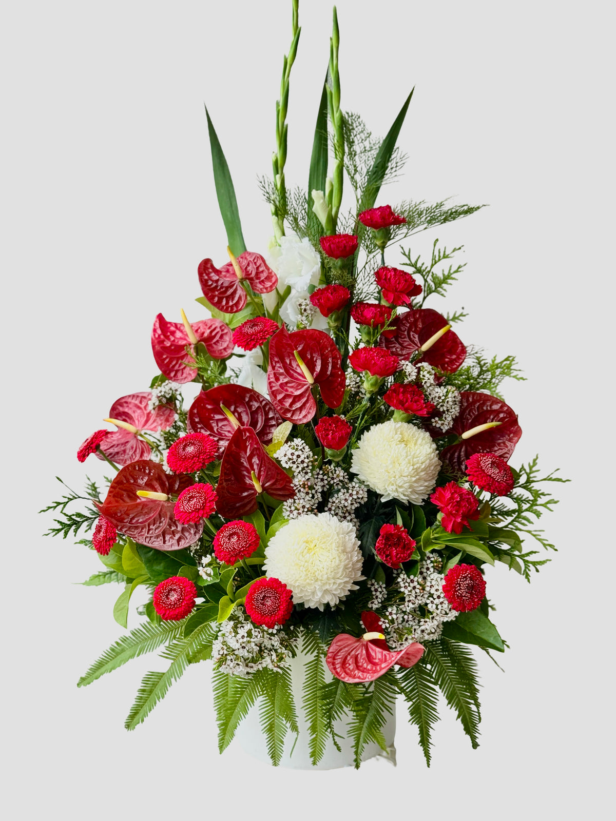 Festive red and white floral arrangement with anthuriums, chrysanthemums and seasonal greenery in a white ceramic pot.