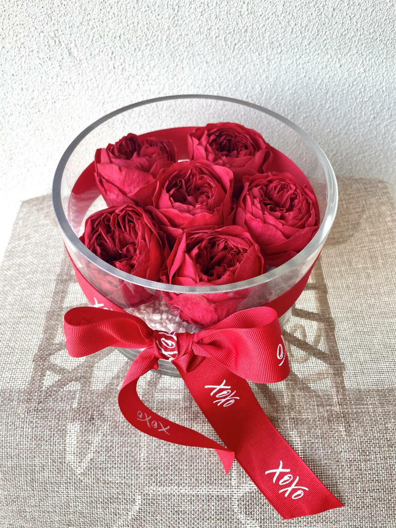 A clear glass vase containing preserved red roses, adorned with a red ribbon and a white fabric lid.