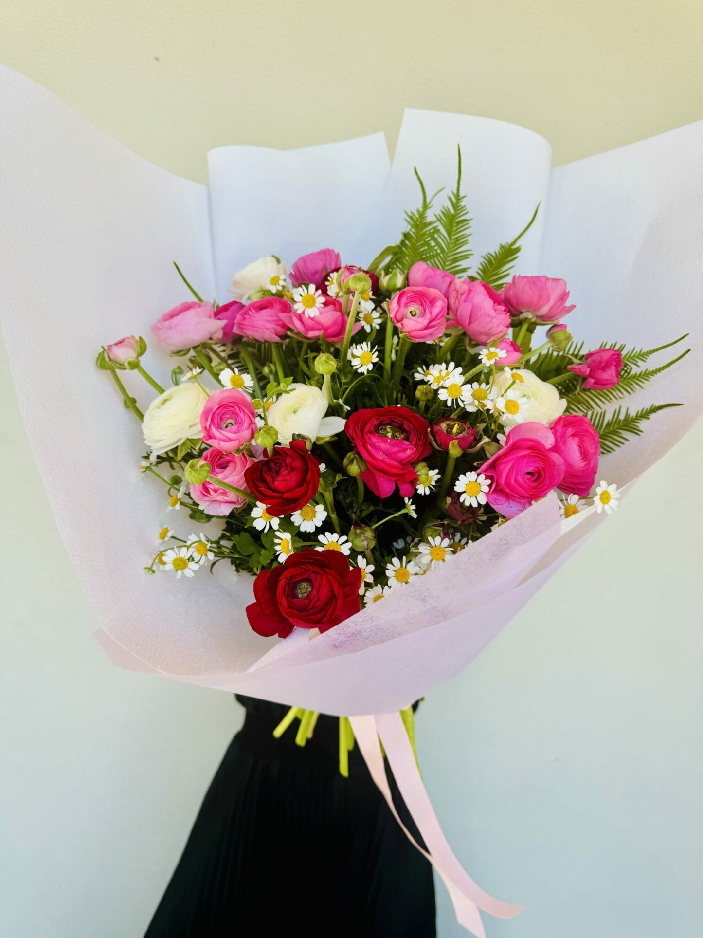 A bouquet of pink, cream, and red ranunculus flowers, arranged and presented in a pink paper bag.