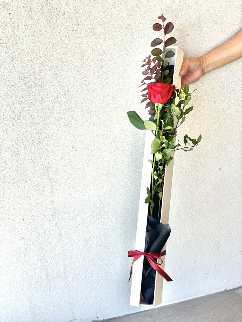 A single long stemmed rose with filler and foliage in black satin like wrap in a white lidded gift box with a satin ribbon.