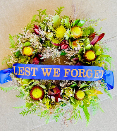 A Anzac Day wreath with a blend of Australian native flora, predominantly greenery, and a blue ribbon with the inscription 'Lest We Forget'.