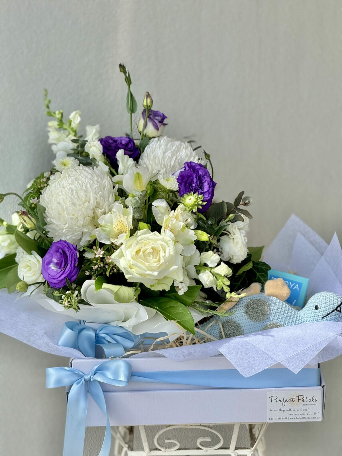 A baby hamper featuring a seasonal floral arrangement in white, mauve, and purple, with a Nana Huchy Dinosaur plush toy and a mini Ecoya candle.