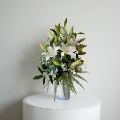 A white ceramic vase filled with white oriental lilies and green foliage, displayed on a white pedestal against a neutral background.