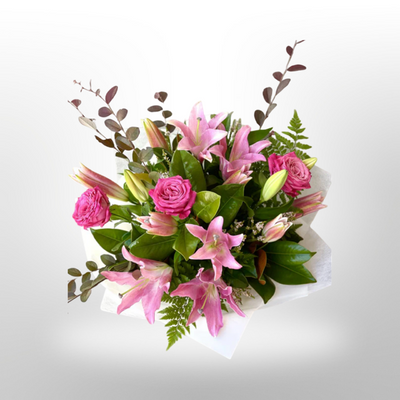 A bouquet of pink oriental lilies and roses with green ferns and other complementary foliage, arranged in a round white basket.