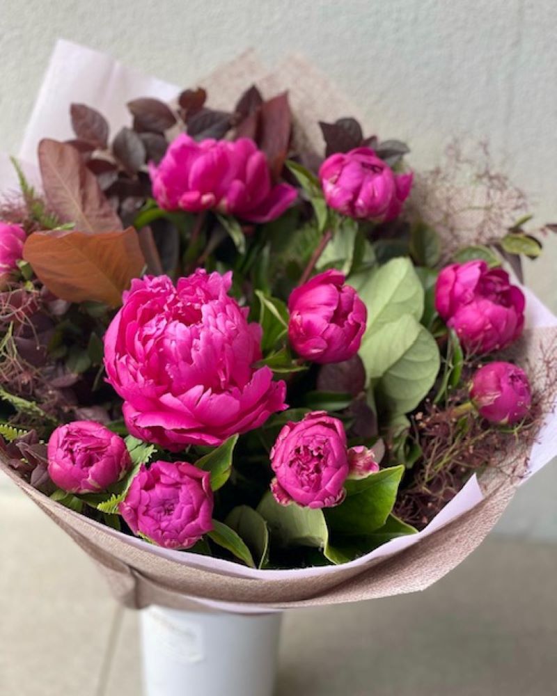 A bouquet of pink peonies with green leaves and brown wrapping paper, placed on a white surface.