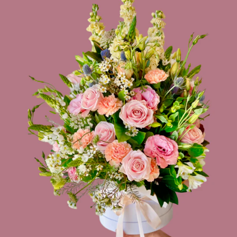 White hat box arrangement of soft pastel blooms including roses, lissianthus, carnations, stocks, sea holly, geraldton wax with lush textural foliage. 