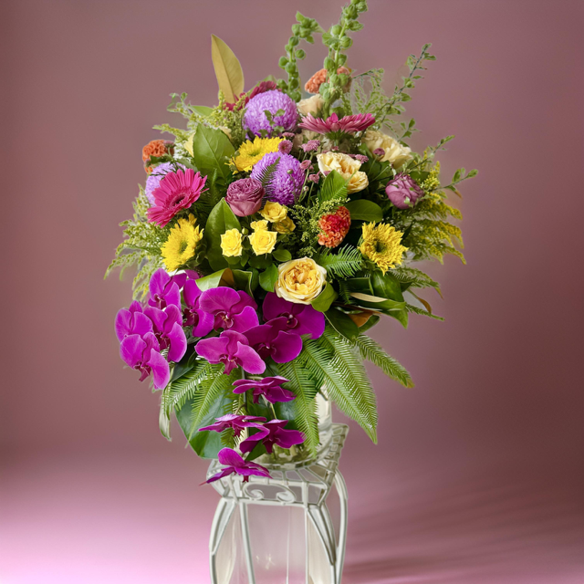 A floral arrangement featuring cascading magenta orchids, sunny yellow roses, gerberas, and textured blooms in shades of lavender and golden hues, complemented by lush greenery.