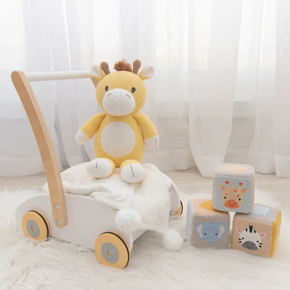 A plush giraffe toy with a yellow body and brown spots, seated on a white cart, with colorful blocks in front.