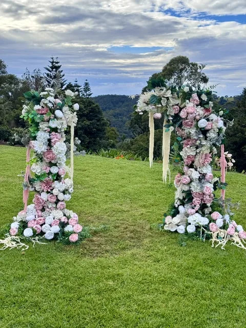 A pair of silk floral pillars in blush and white flanking a lawn ceremony site, with hills and bushland behind