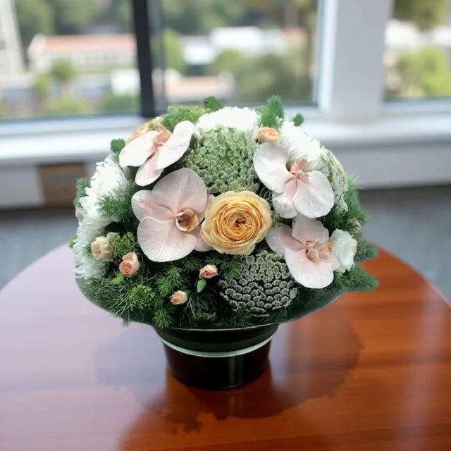 A floral centerpiece with a round shape, featuring a mix of white disbuds,  peach roses, white and peach phalaenopsis orchids and green flowers and lush green foliage, placed on a table.
